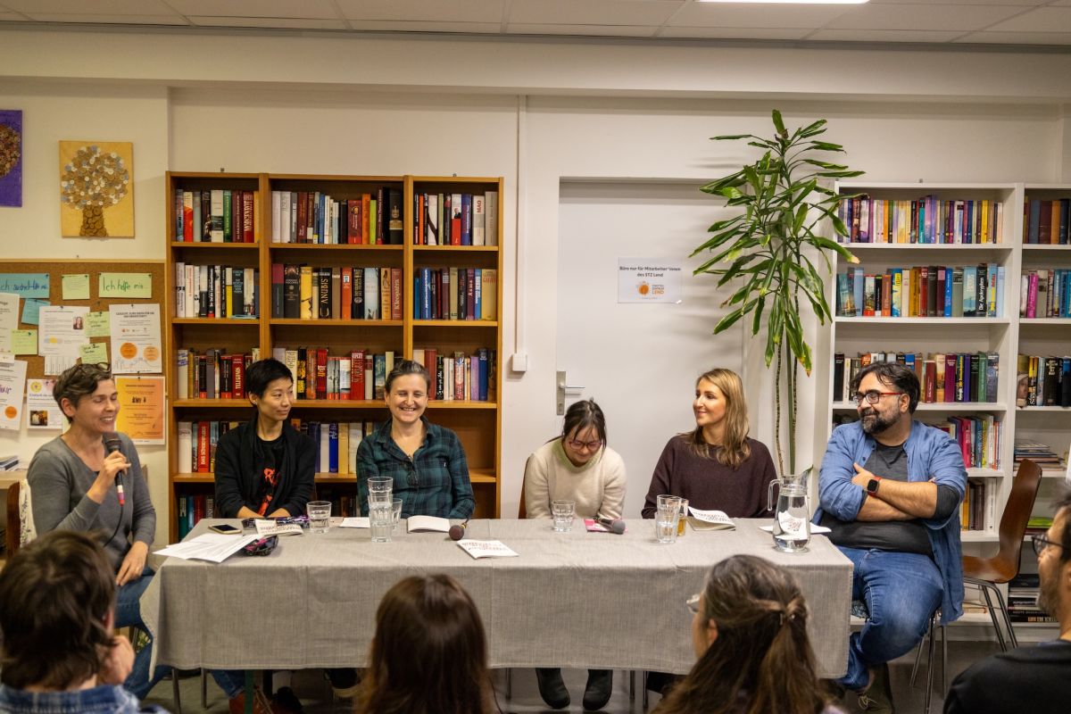 Lachende Teilnehmer:innen einer Podiumsdiskussion vor zwei Bücherregalen, eine Person hält ein Mikrofon.
