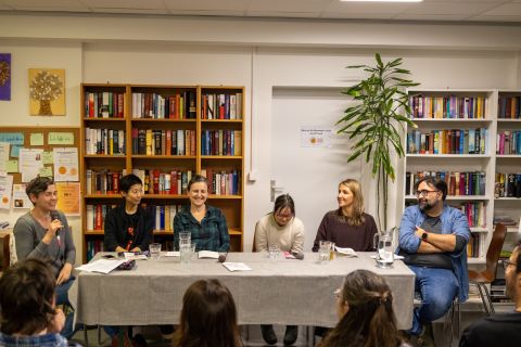 Lachende Teilnehmer:innen einer Podiumsdiskussion vor zwei Bücherregalen, eine Person hält ein Mikrofon.